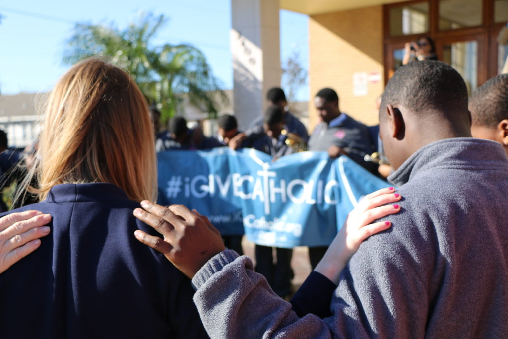 Catholic school students in a circle, with their hands on each others' shoulders. They look like they're pausing for prayer. There is an "IGiveCatholic" sign in the background