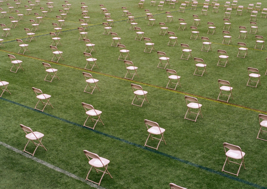 Rows of chairs outside on a green field of grass, separately 6 feet in all directions. The chairs are all empty.