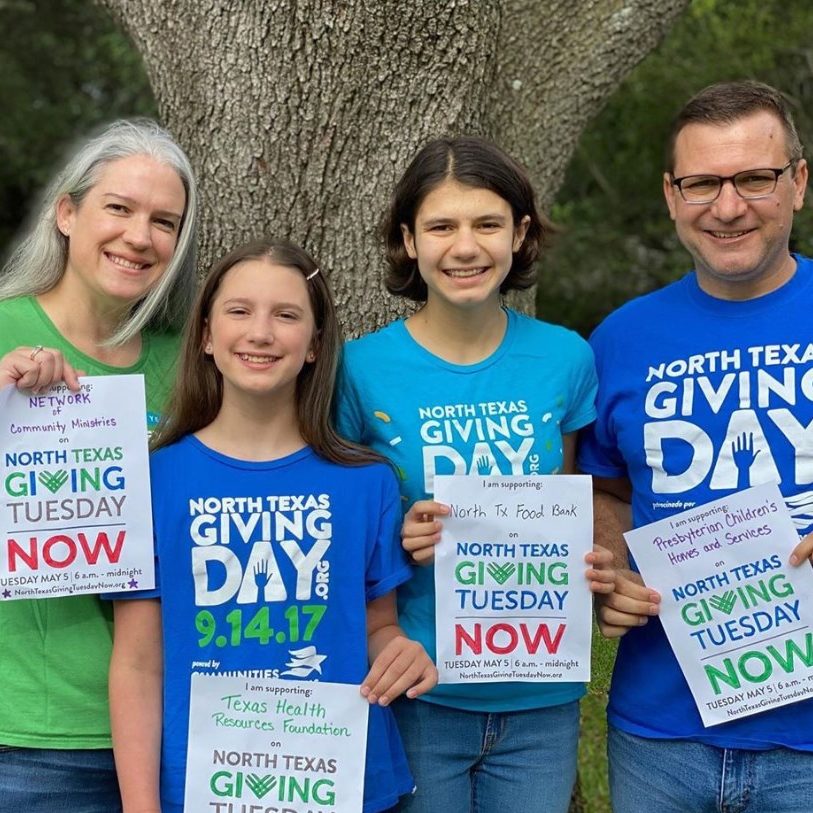 A family of 4 standing in front of a tree, smiling and holding signs that say " North Texas Giving Tuesday Now"
