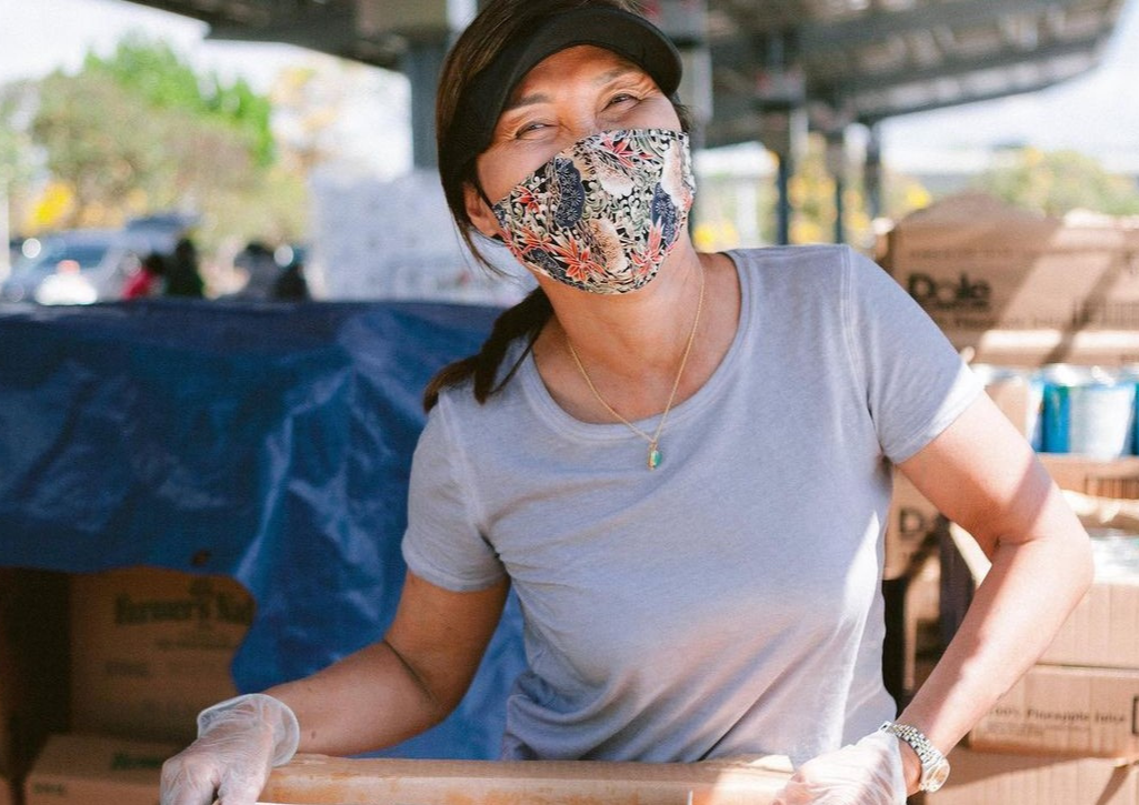 Asian woman wearing a mask and a black visor. She's holding a box and is volunteering at a food distribution event
