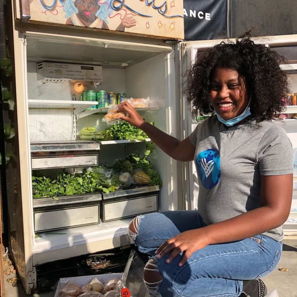 A Black girl in a GivingTuesday tshirt posing for a photo while stocking her local community refridgerator with fresh vegetables