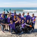 Harrahs Casino employees wearing purple shirts and posing with open arms on a beach that they've just cleaned up on GivingTuesday