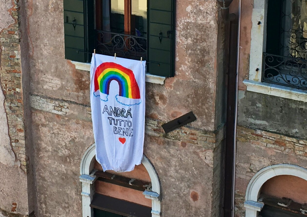 A homemade sign hanging out of a window of a building in Italy. There's a rainbow on it and it says "Andra Tutto Bene"