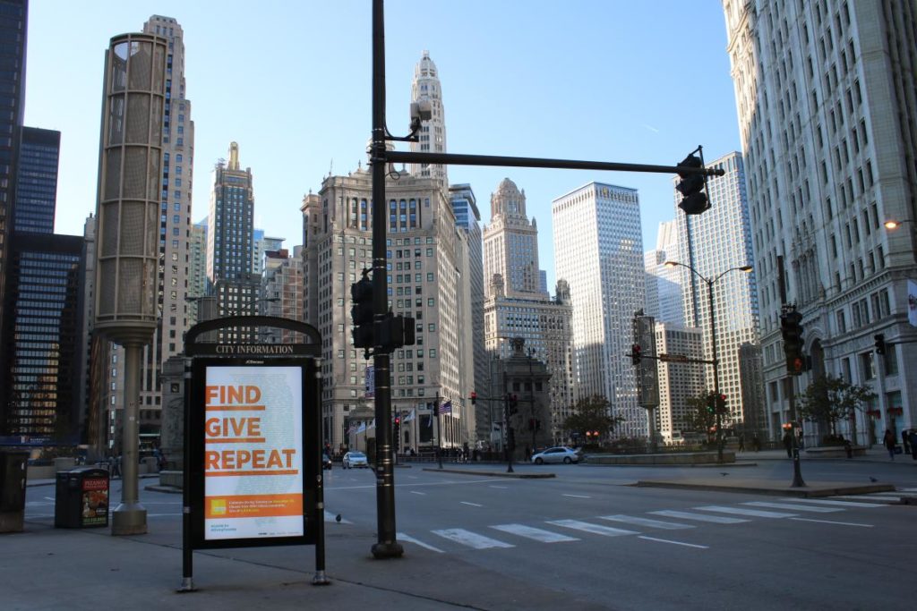 A street corner in Chicago with a information board that says " Find Give Repeat" on it.