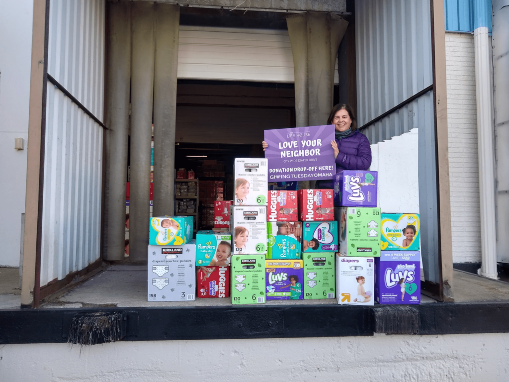 a citywide diaper drive in Omaha - a woman holds a sign that says "GivingTuesdayOmaha" and she's standing next to a stack of a dozen boxes of various diaper brands stacked in a pyramid shape