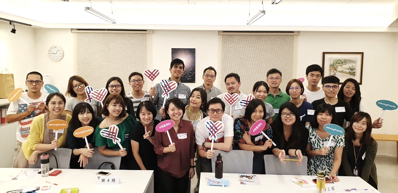 Photo of  a group of people holding heart signs in a room with white walls and tables during GivingTuesday Taiwan meetup