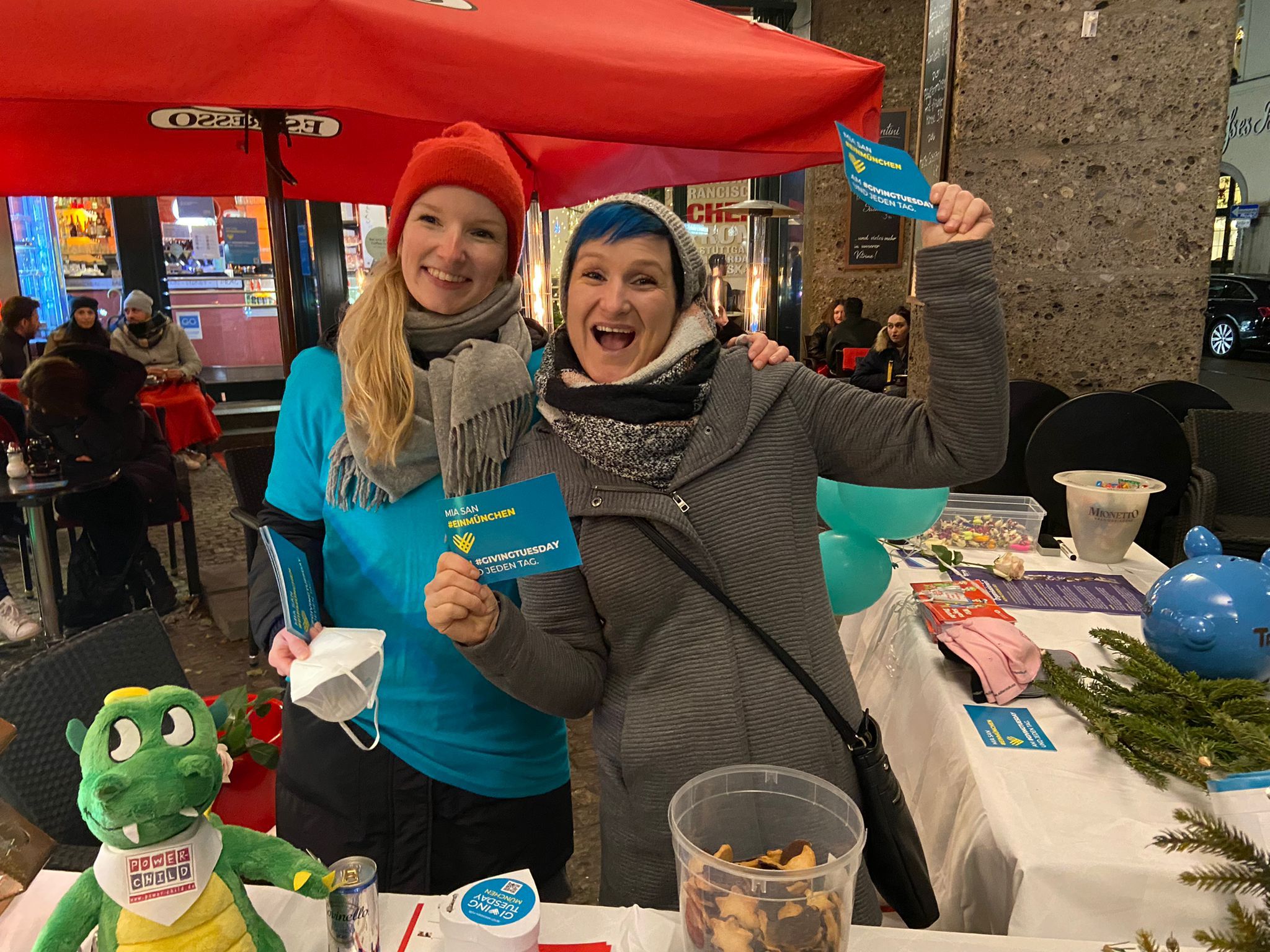 Two women smiling and wearing winter clothes at an outdoor festival in Germany. They're stationed at the GivingTuesday table and are spreading the word, sharing games and prizes, and inspiring others to give back in honor of GivingTuesday