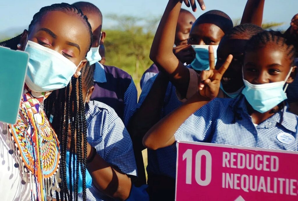 An African woman wearing a mask taking a selfie with African kids. One of the kids is holding a pink sign about SDG 10 "reduced inequality"