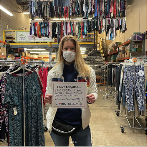 A woman holding an unselfie in front of racks of clothing at a women's empowerment nonprofit. She's holding a sign that says "I give because I am grateful for what I have and want others to thrive."