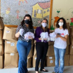 Brazilian women wearing masks and standing in front of stacks of boxes: goods they've collected for covid prevention