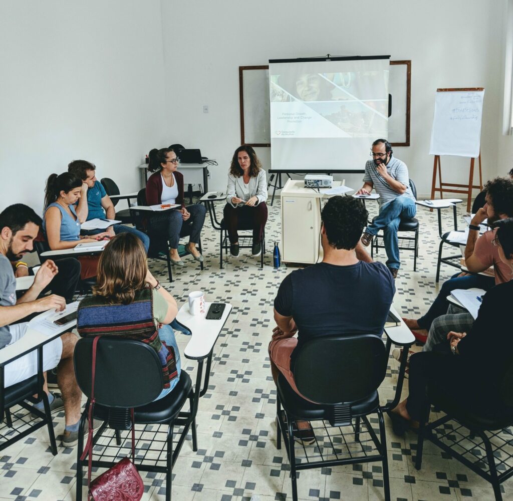 A group of people sitting in chairs in a circle (pre-pandemic), listening to a lecture and getting ready to brainstorm