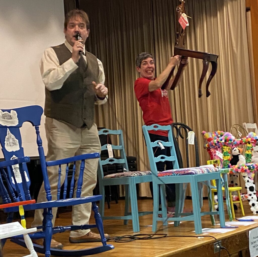 Two men on a stage holding up painted chairs. THey're auctioning them off for a community dinner