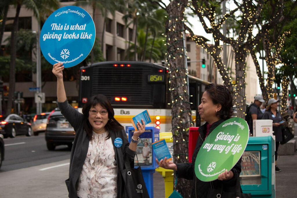 a woman holding up a sign that says "this is what giving looks like". She's on a street with a bus near her.